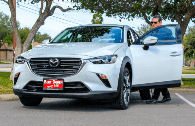 A black Mazda hatchback is parked on the side of the road, and a man is walking in front of it.