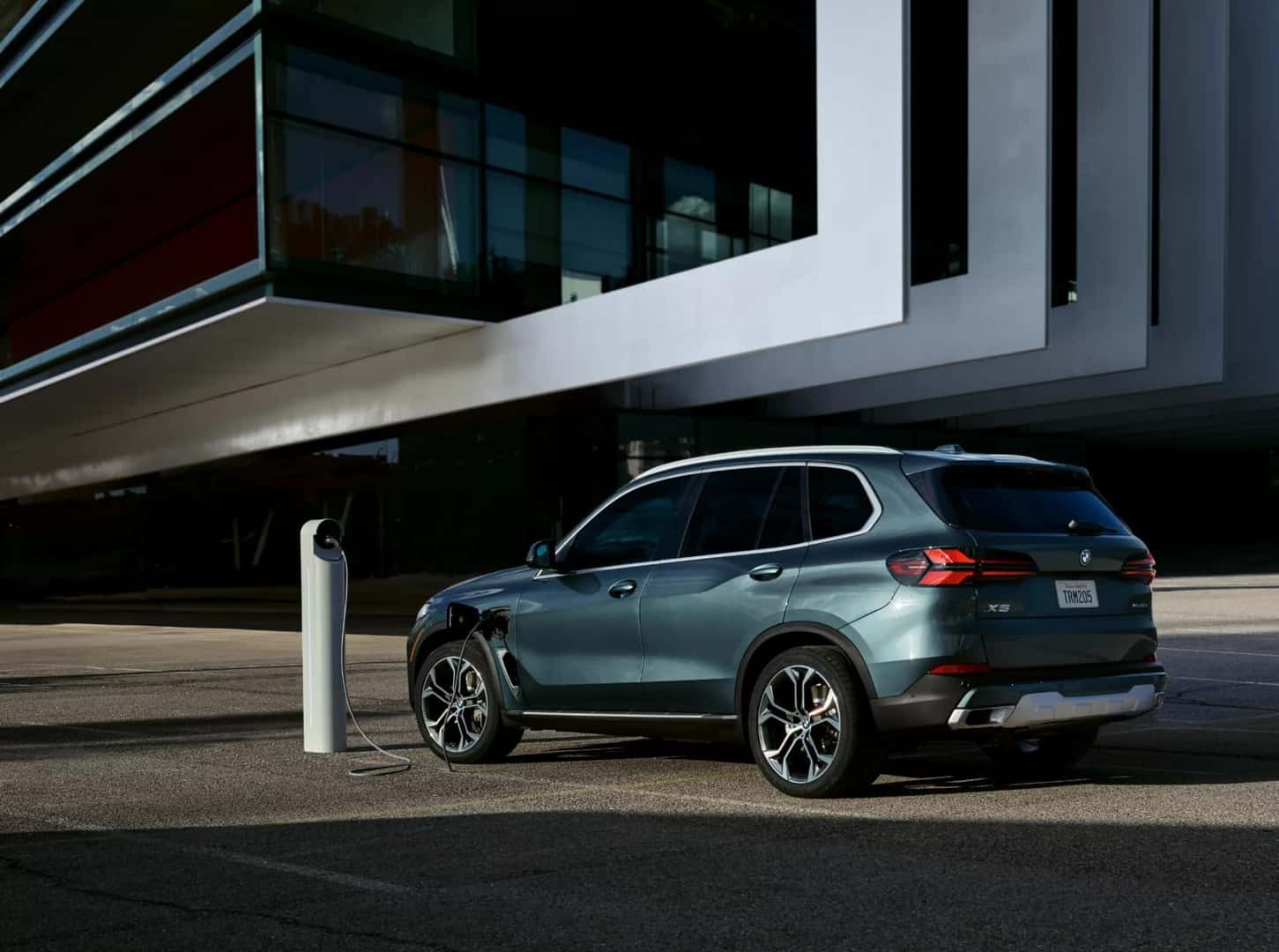 A BMW X5 beside a modern building, plugged into a charging post