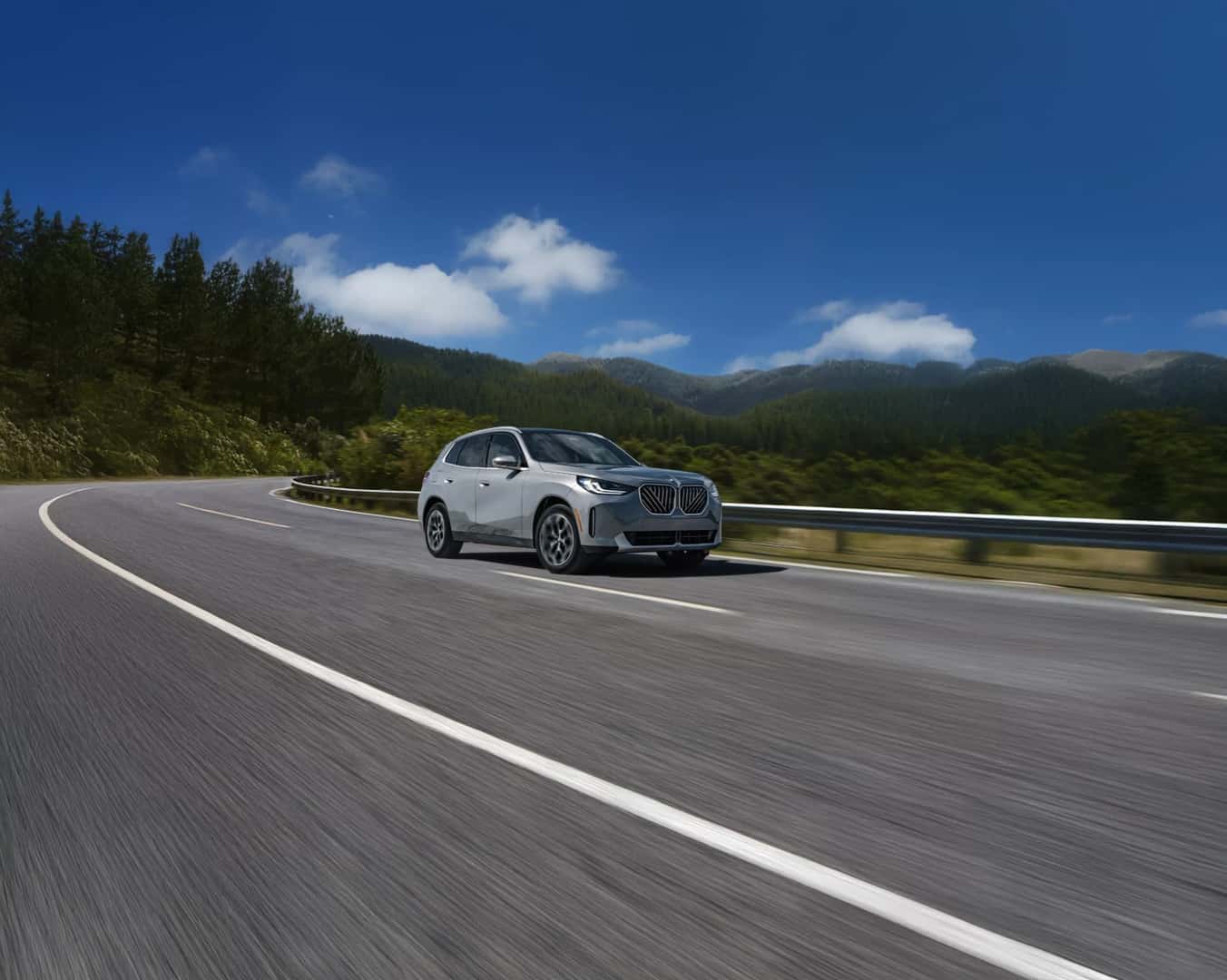 A silver BMW X3 driving on a mountain road lined with pine forest