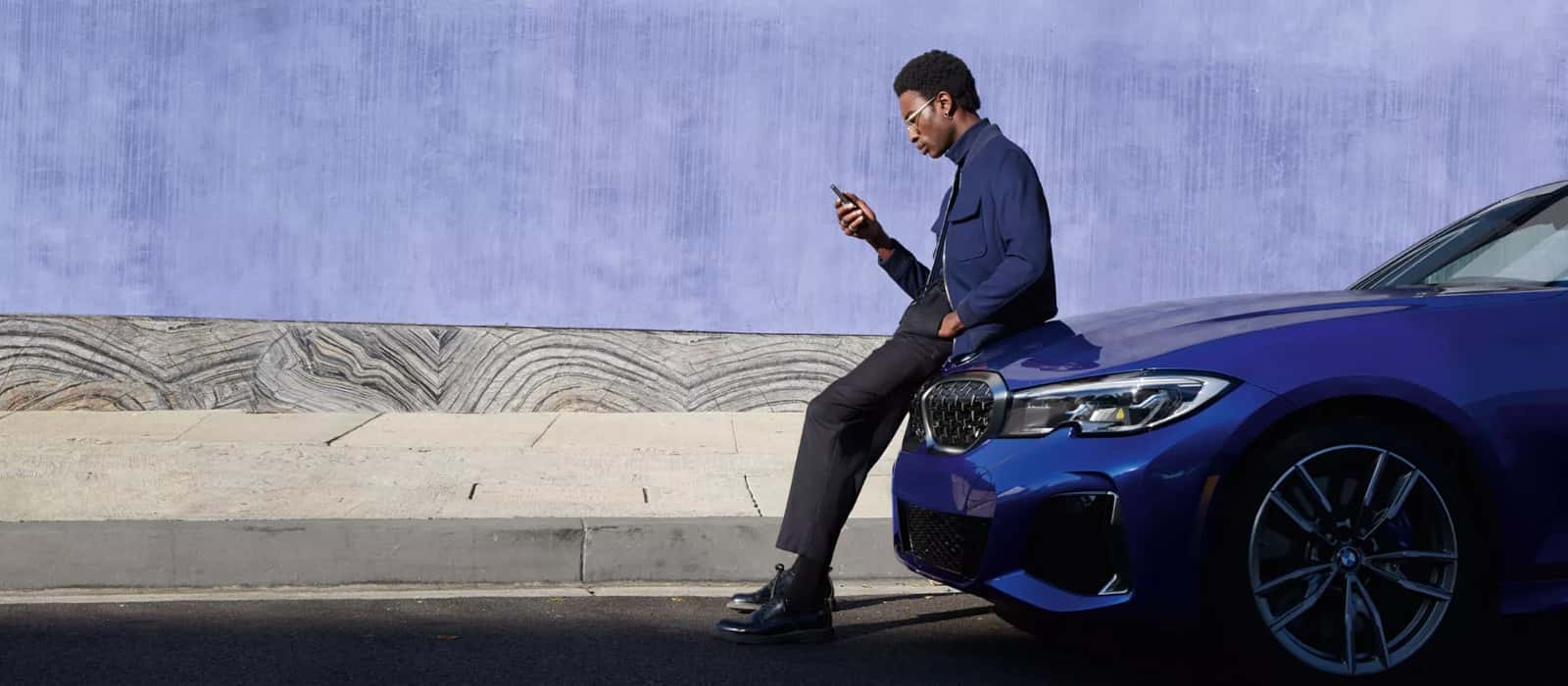 A man sitting against a blue BMW, looking at his phone