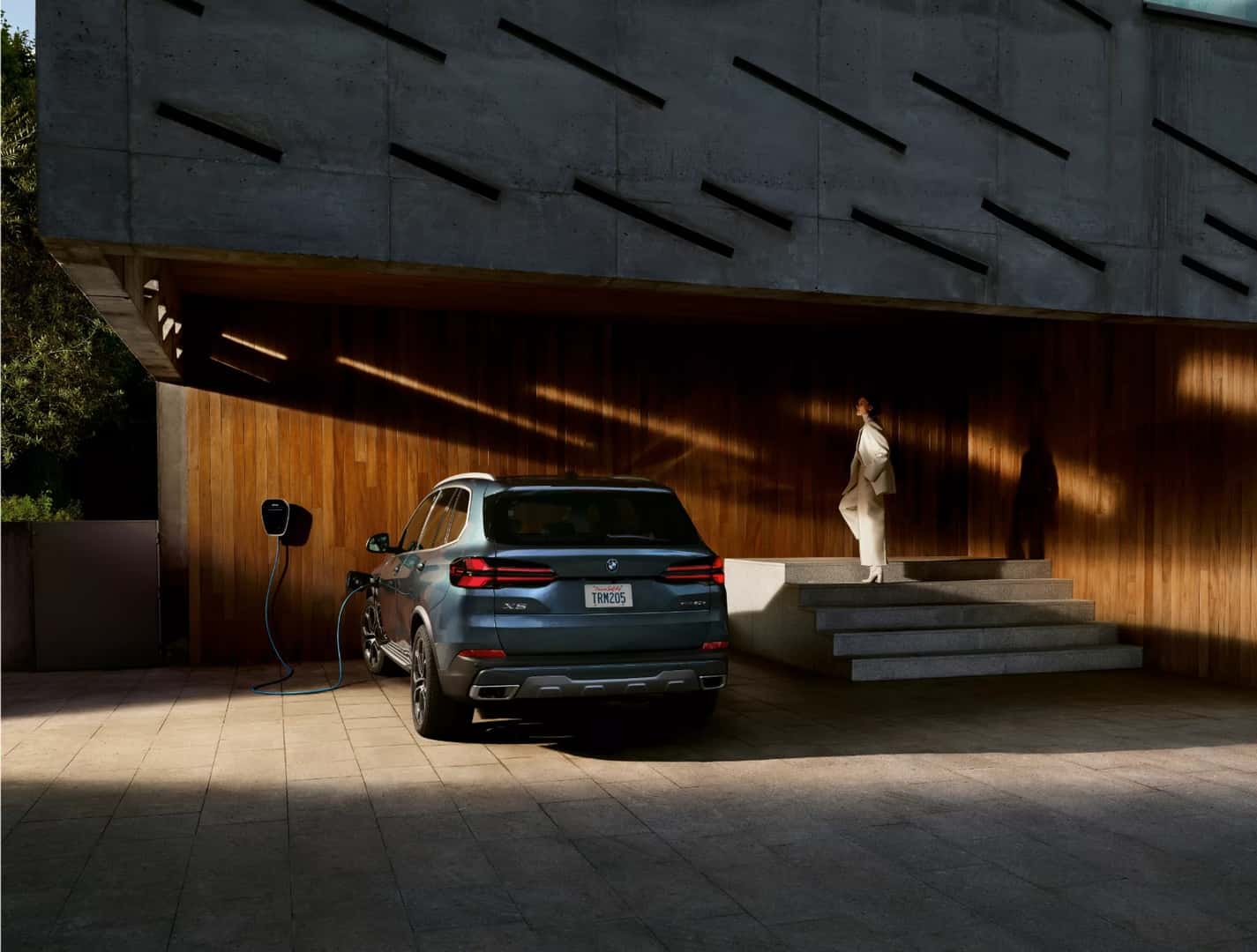A BMW X5 parked and charging under the overhang of a modern building, seen from behind
