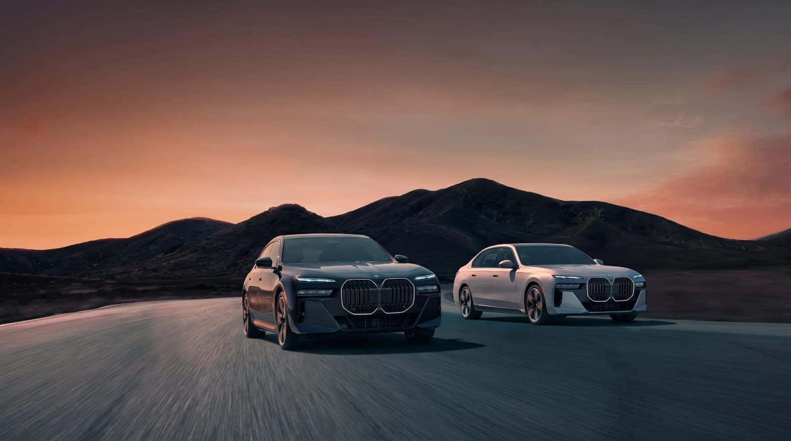 Two BMW 7 Series sedans driving side by side on a mountain road at dusk