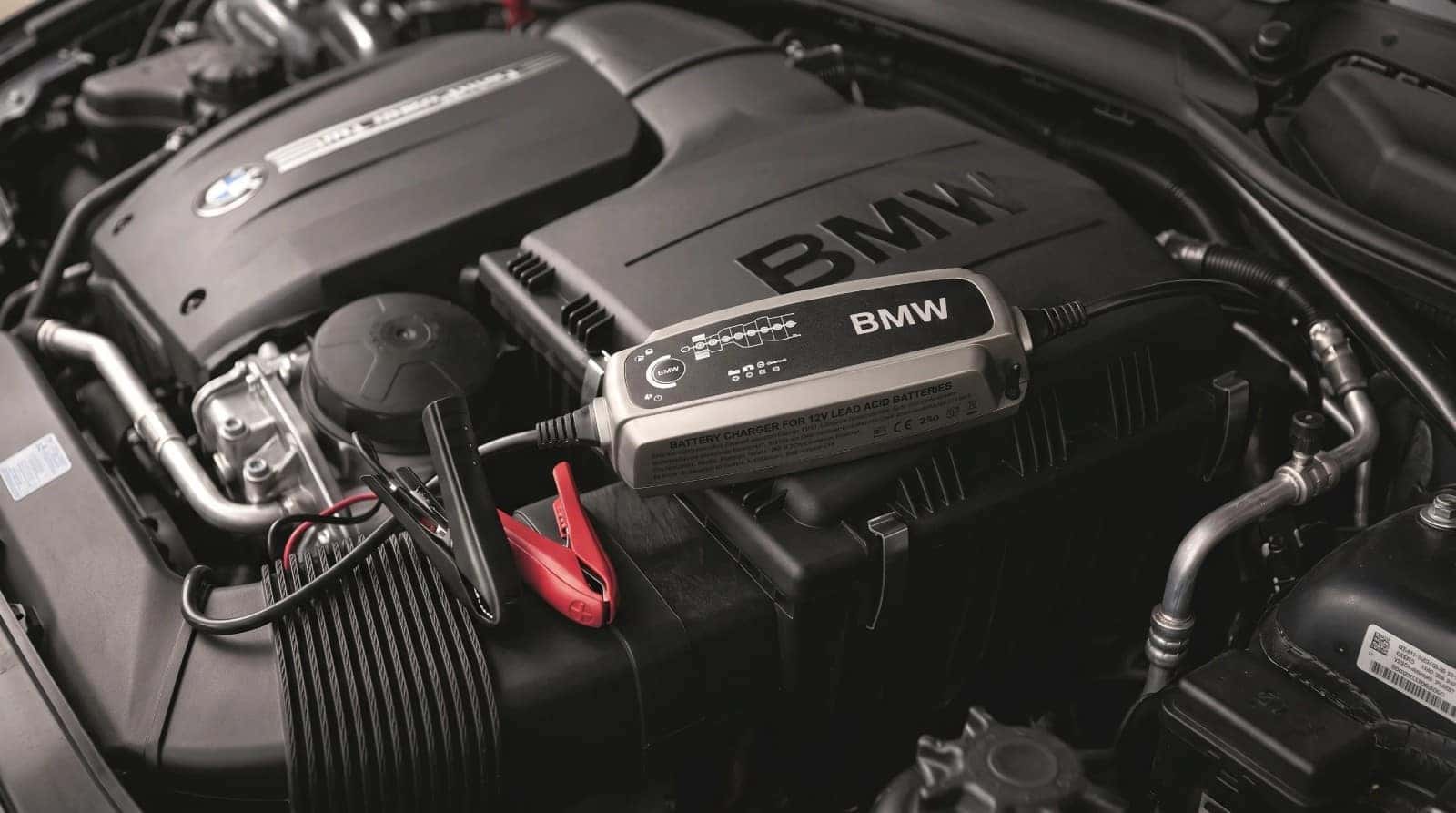 A BMW battery charger resting on the engine cover with its clamps on the battery