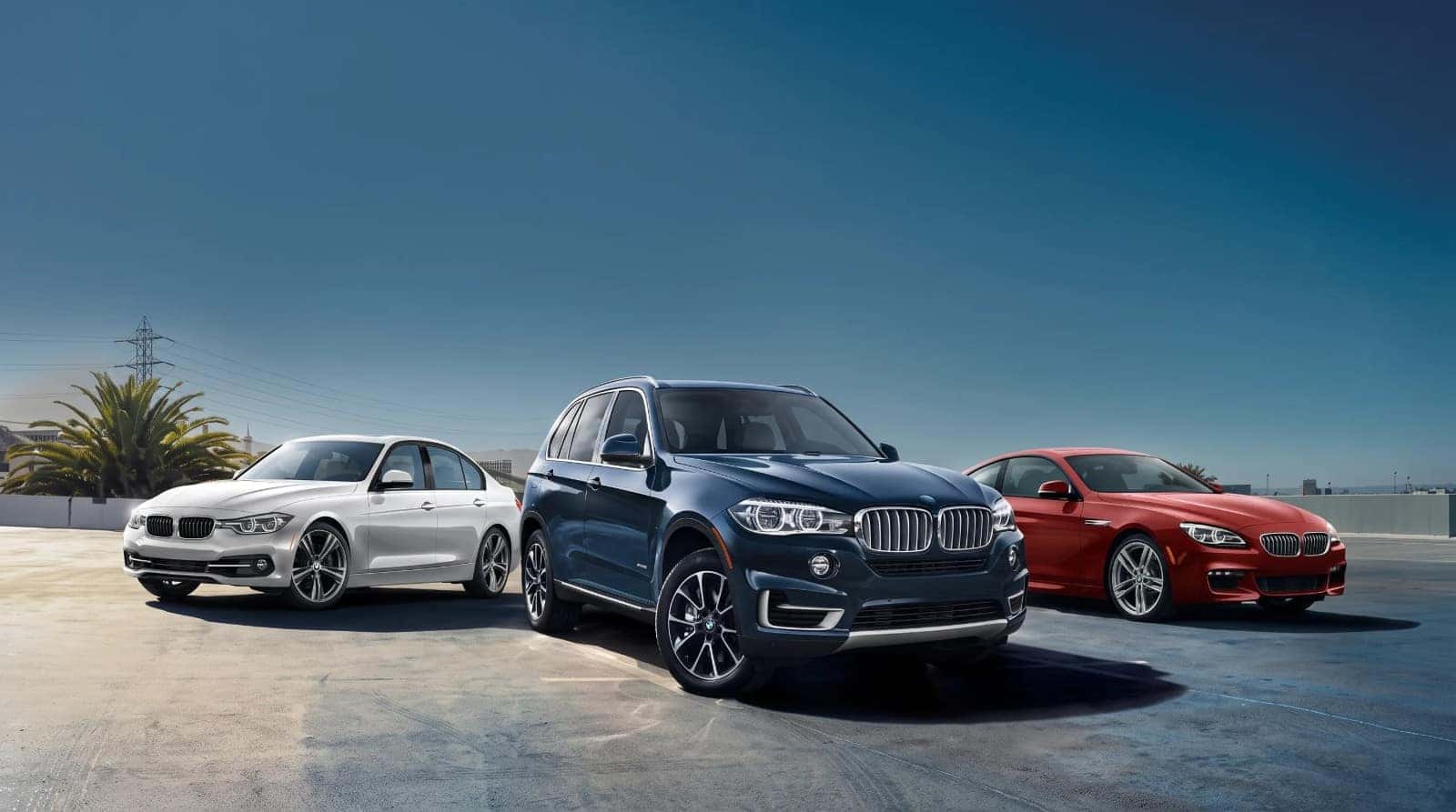 A white BMW 3 Series, a dark blue X5 and a red 6 Series parked side by side under a clear sky
