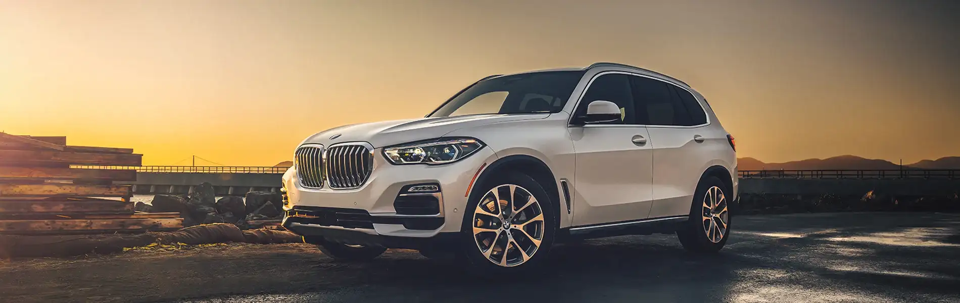 A white BMW X5 parked on a shoreline road at sunset, front three-quarter view
