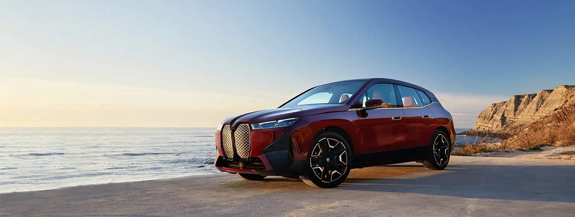 A red BMW iX parked on a coastal road beside the ocean, with cliffs behind it