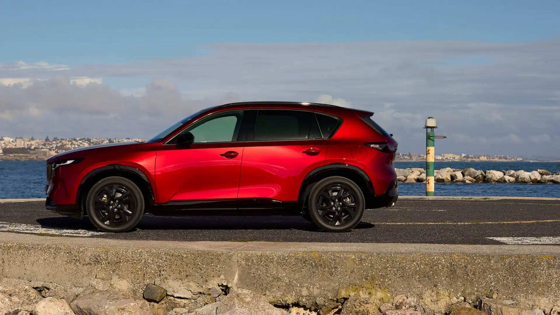 2026 Mazda CX-5 red SUV side profile parked beside waterfront rocks with black wheels and skyline
