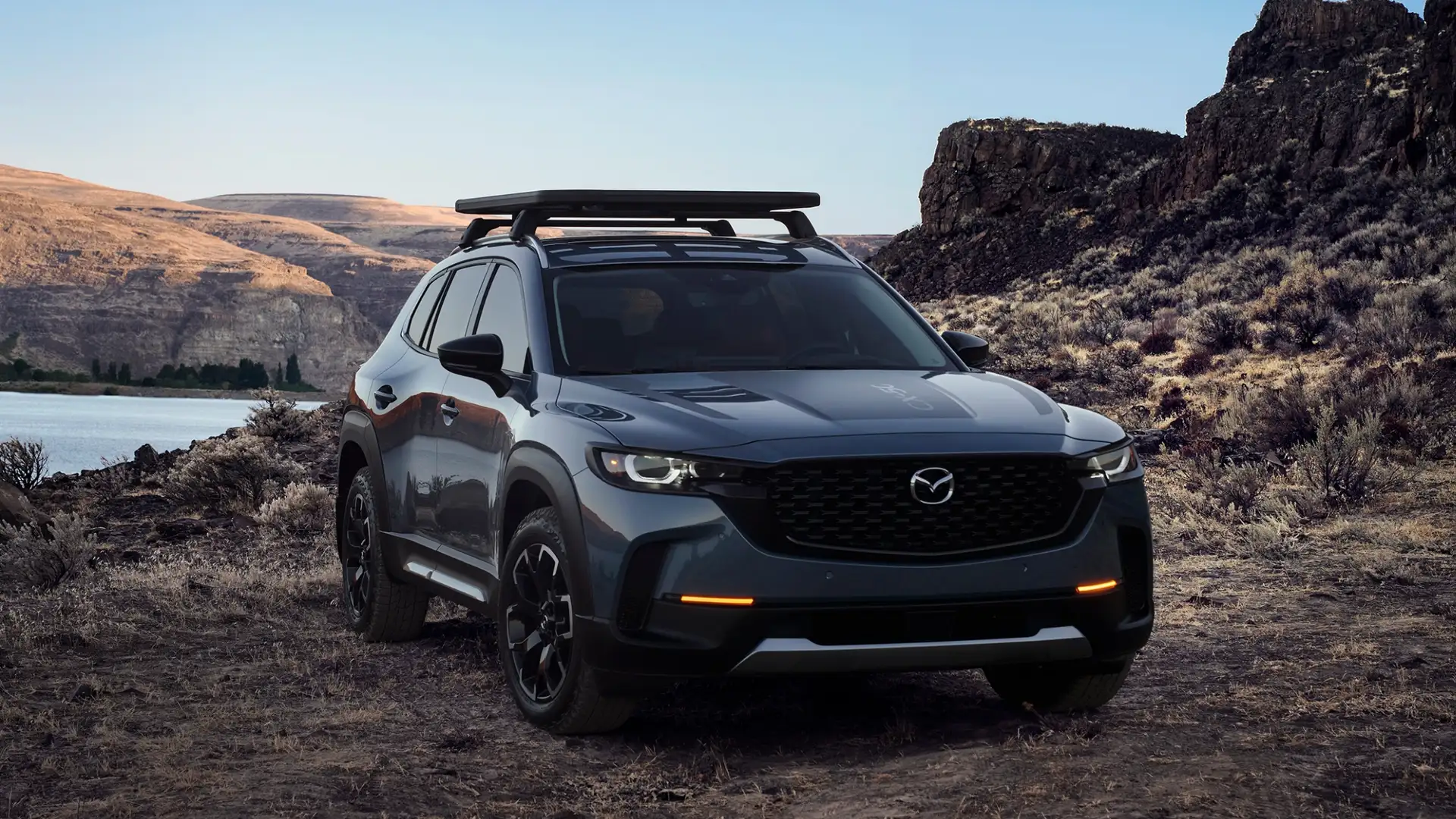 2026 Mazda CX-50 gray SUV with roof platform parked by canyon lake and rocky desert terrain