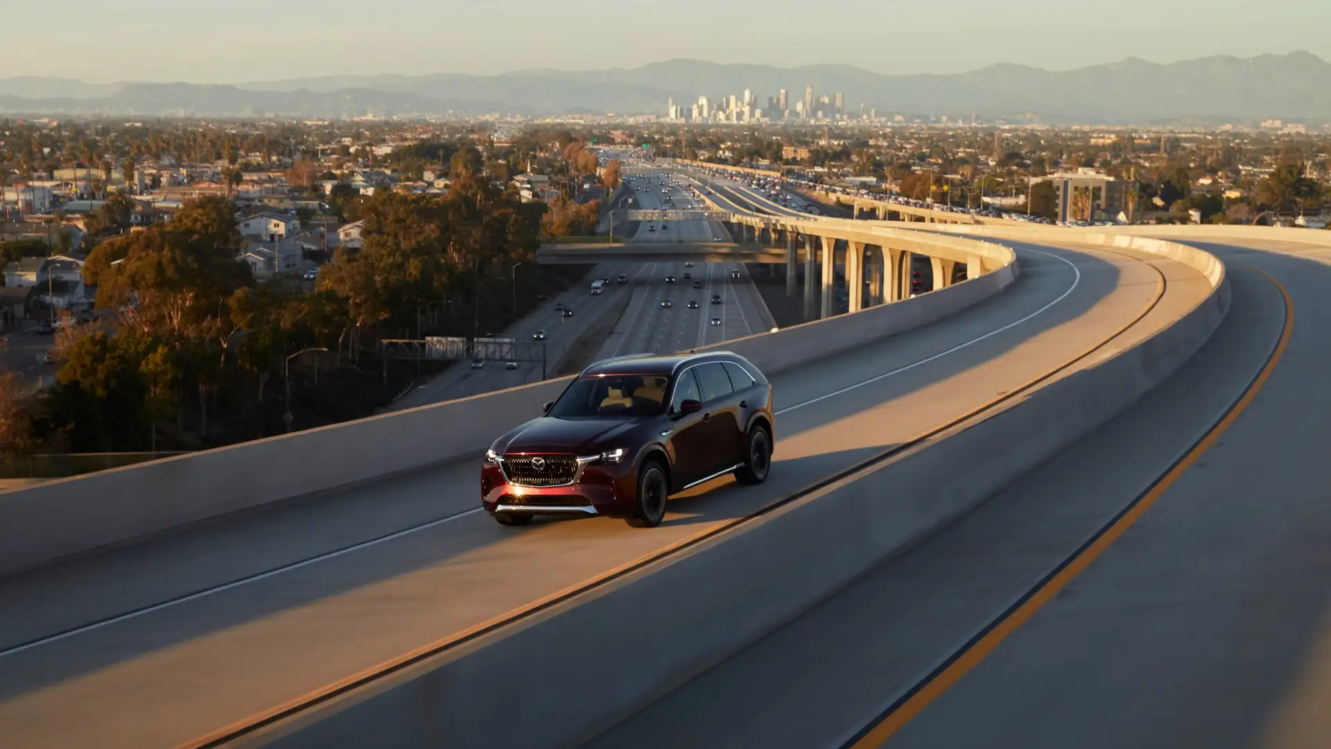 2026 Mazda CX-90 red SUV driving on elevated highway ramp with city skyline in the distance