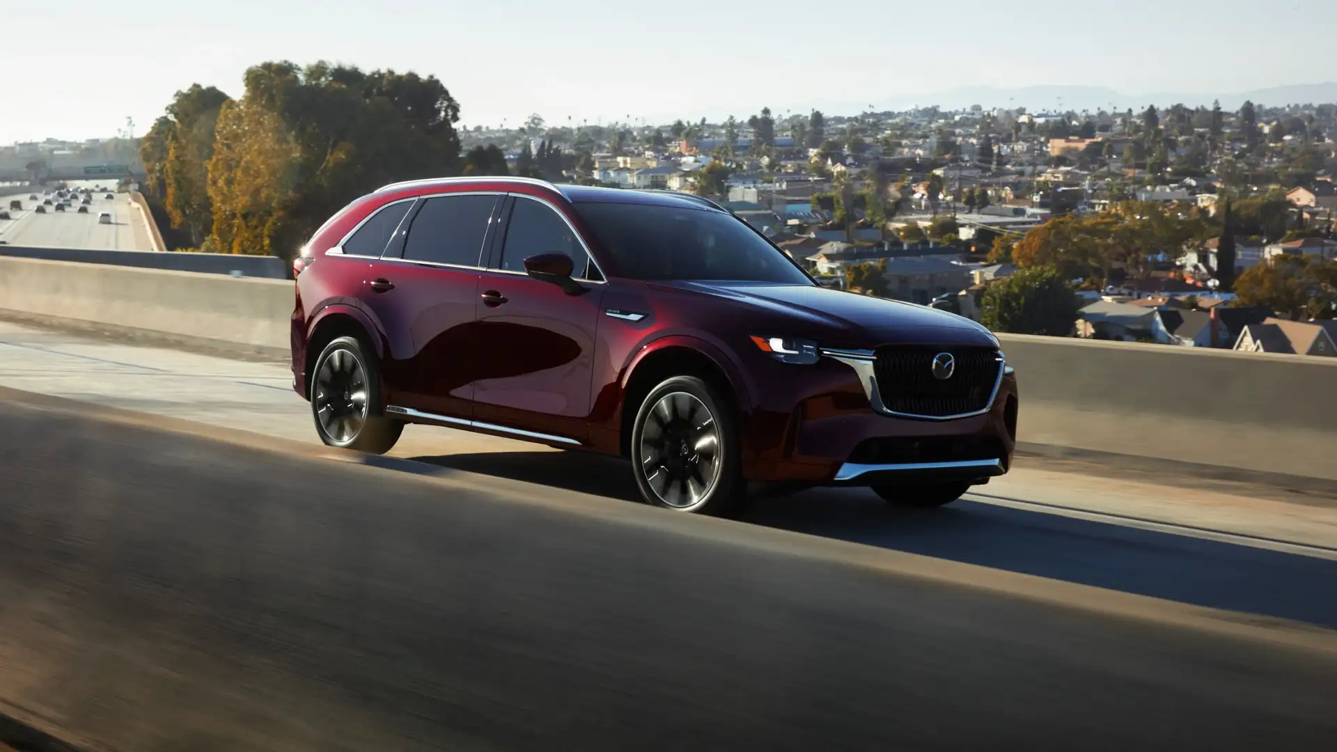 2026 Mazda CX-90 red SUV driving on elevated highway with city homes and mountains in background