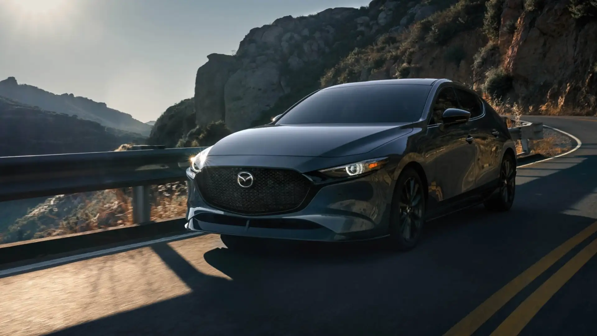 2026 Mazda3 Hatchback dark gray car driving on winding mountain road with rocky cliffs at sunset