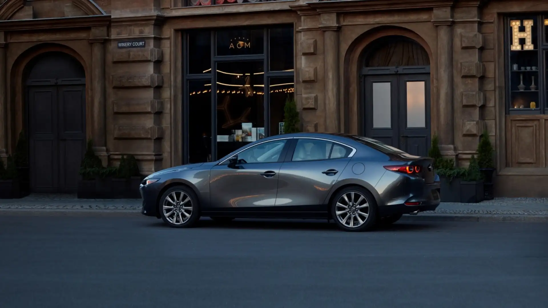 2026 Mazda3 Sedan gray side profile parked along a city street by illuminated storefronts at dusk