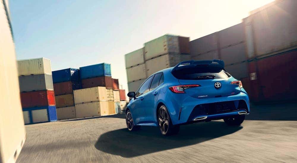 A blue 2020 Toyota Corolla Hatchback is driving through a shipping container storage area.