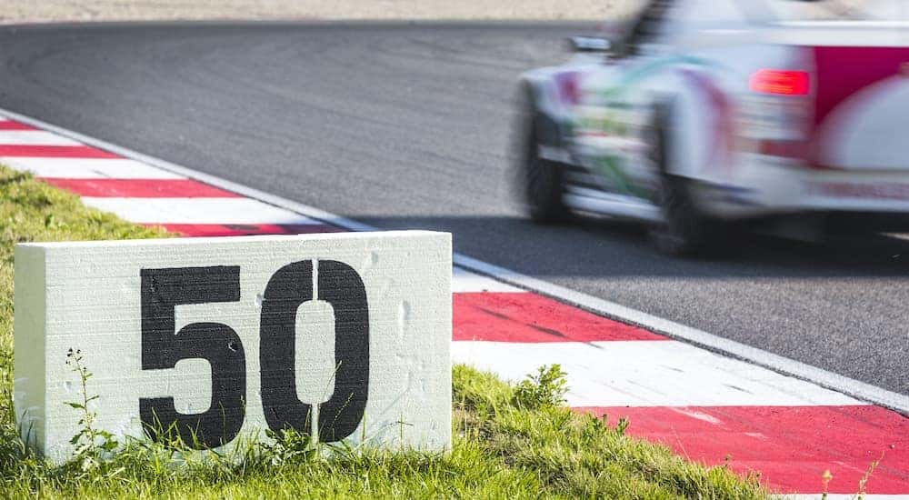 A close up shows a distance sign on the corner of a race track with a race car in the background.