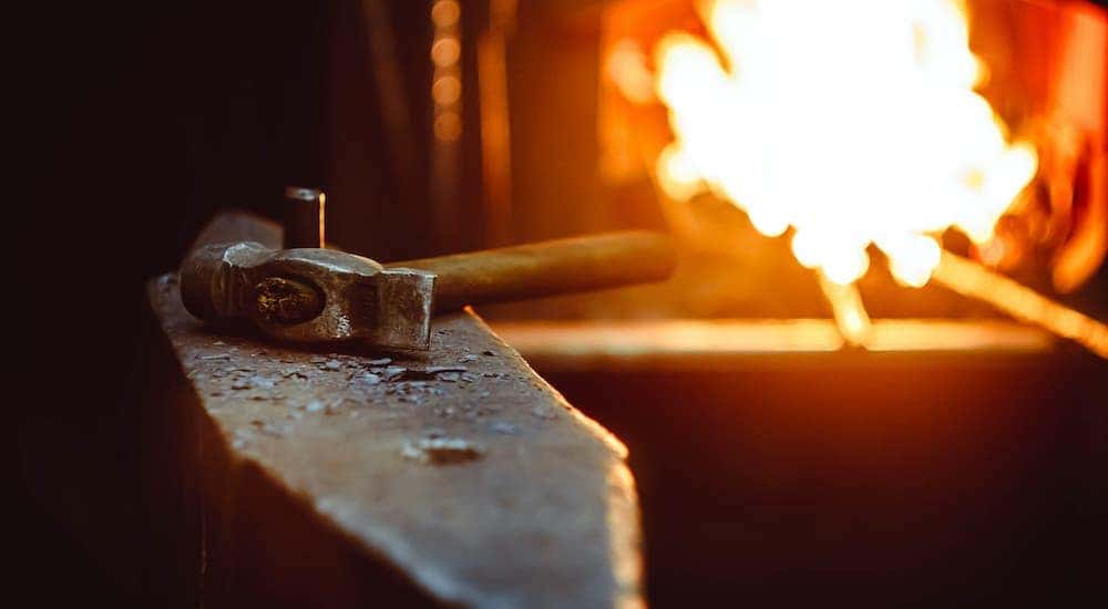 A close up shows a blacksmith hammer sitting on an anvil with a fire in the background.
