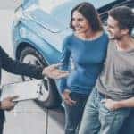 A salesman is talking to a couple while they lean against a blue car at a used car dealership in Defiance, OH.