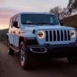 A white 2020 Jeep Wrangler Unlimited is driving on a dirt road at sunset.