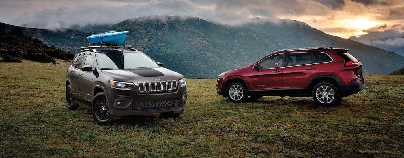A red and a gray 2020 Jeep Cherokee with a kayak on the roof racks are parked in front of cloudy mountains at sunset.