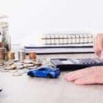 Hand using a calculator, a change jar, loose cash, and a blue toy car