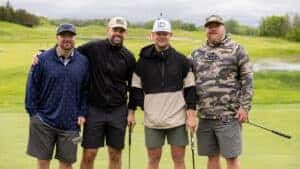 Four men standing together on a golf field holding golf clubs. 