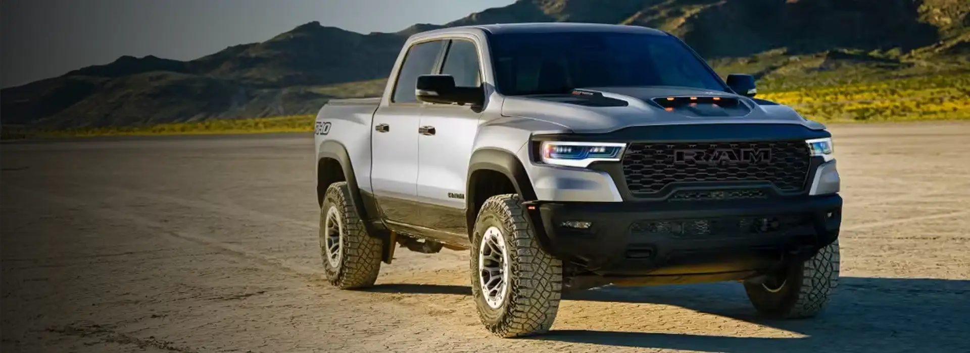 Silver 2027 RAM 1500 TRX pickup with black fender flares parked on a dry desert flat below rocky green mountains